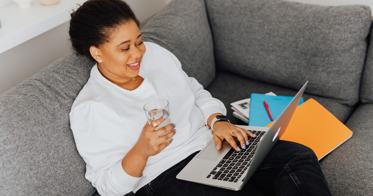 Writer sitting on a couch with glass of water typing on a laptop