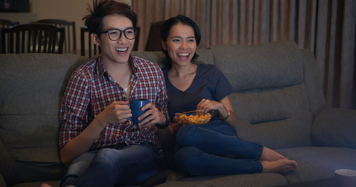 Two smiling people with drink and snack sitting together on couch watching television