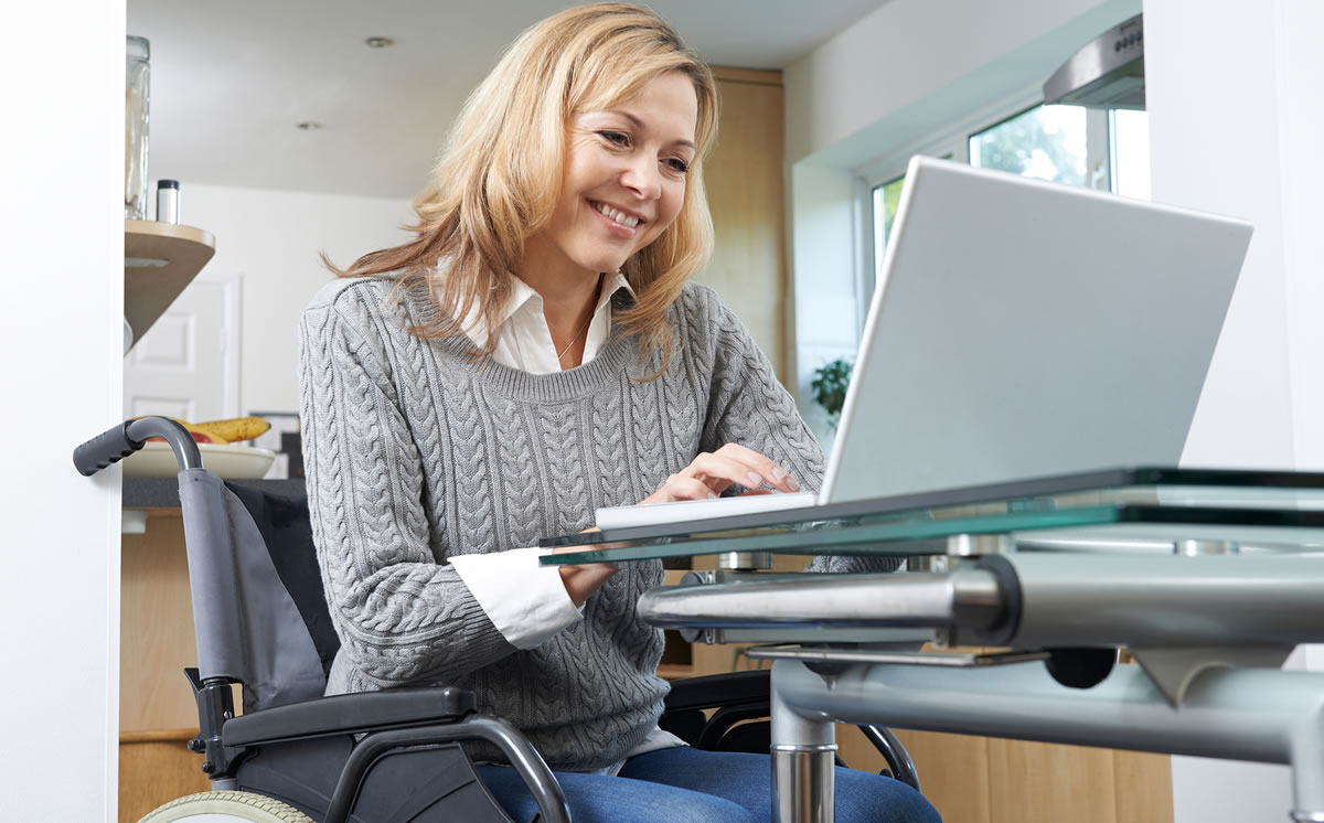 Smiling woman in wheelchair working on laptop at desk at home