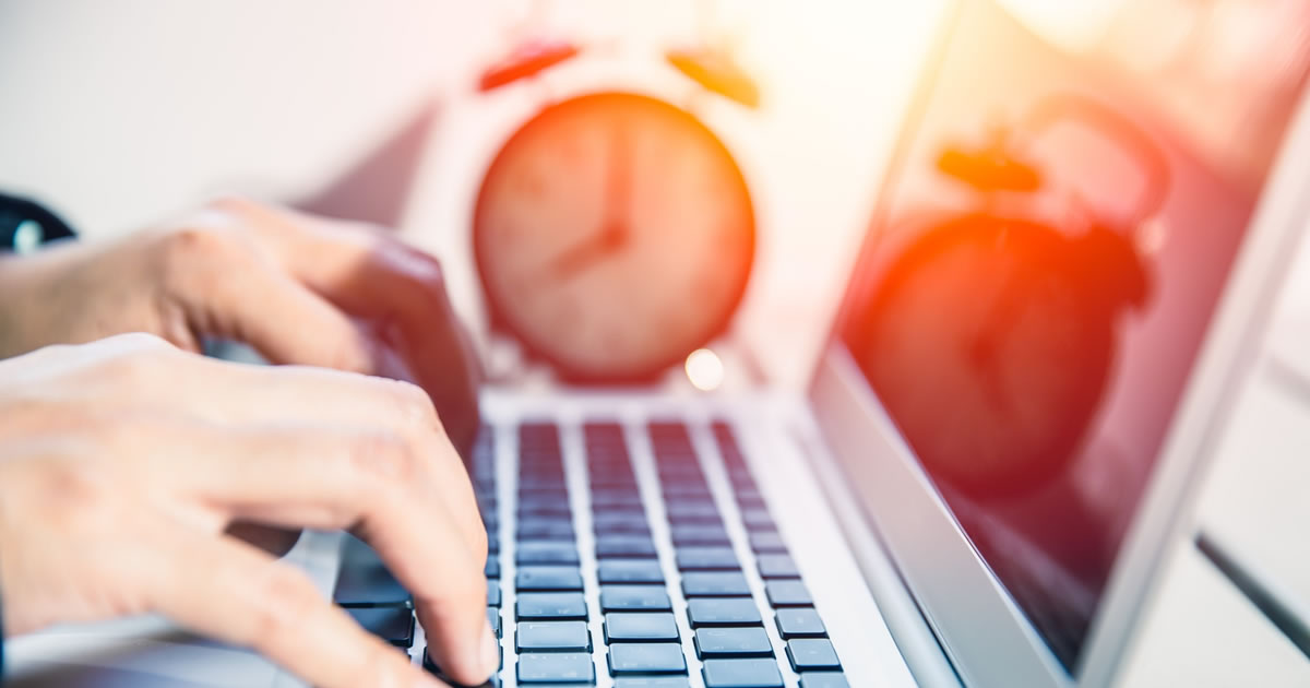 Closeup of writer typing on laptop with clock on desk