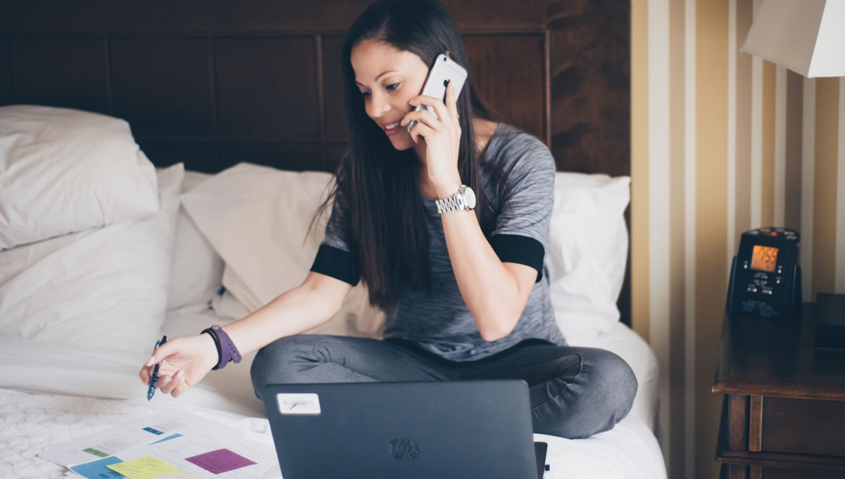 Smiling woman sitting on a bed holding a pen and talking on a mobile phone while looking at an open laptop and papers in front of her