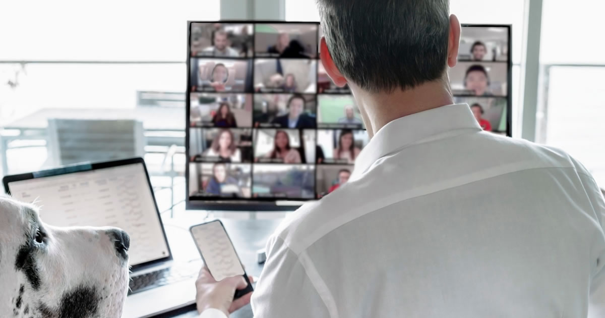 Rear view of a man and a dog sitting at a desk viewing a group video chat screen