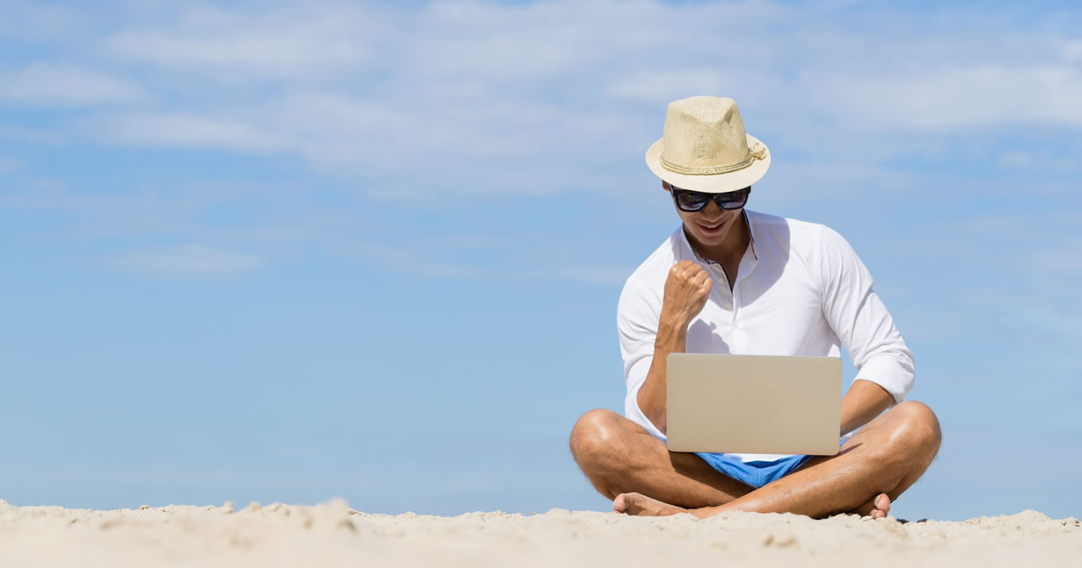Young man writing on his laptop while sitting on a beach with a blue sky background