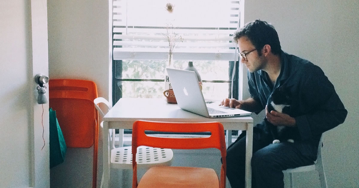 Man wearing pajamas writes on laptop at kitchen table while petting cat in his lap