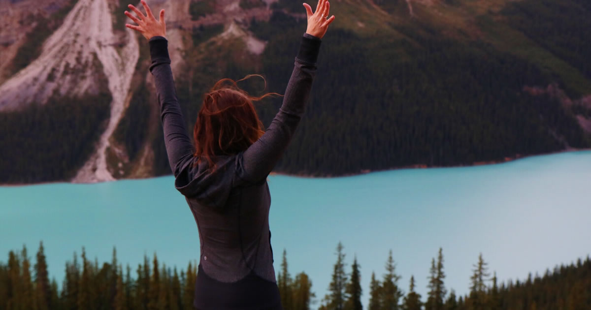 Woman facing away from camera standing outside in front of mountains and water with arms raised in air