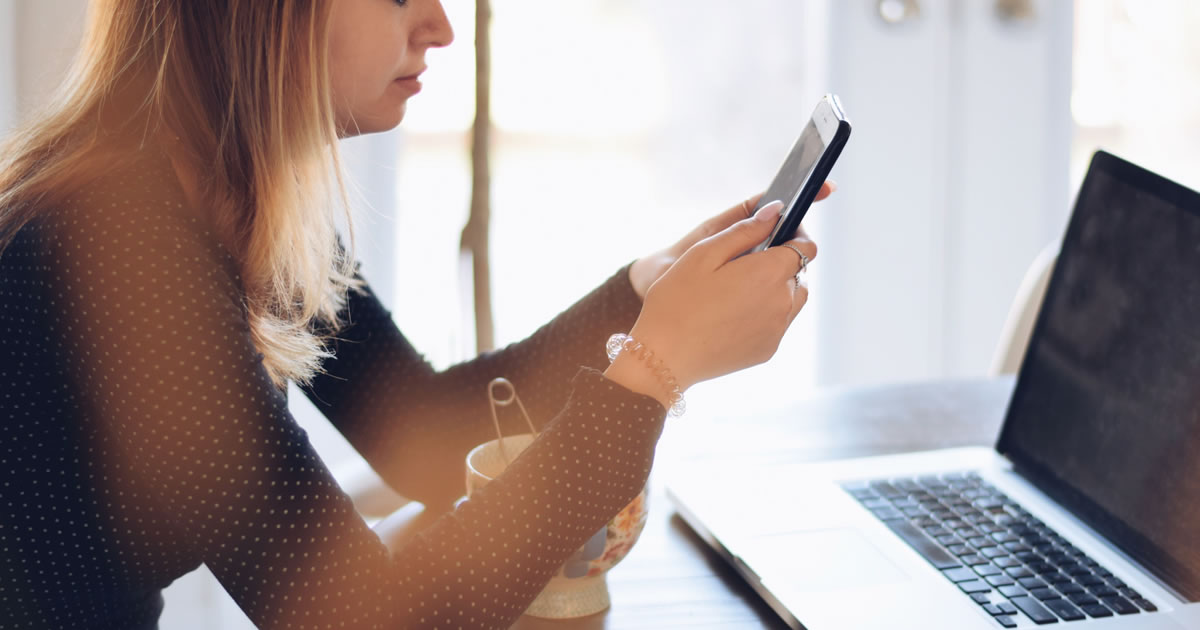 Female looking at phone, sitting at a table with open laptop