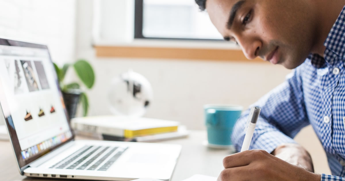 Man writing in a notebook in front of a laptop sitting on desk