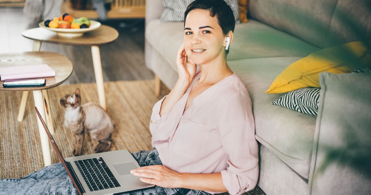 Smiling businesswoman sitting on floor in front of couch next to cat at home while using laptop and wireless ear phones