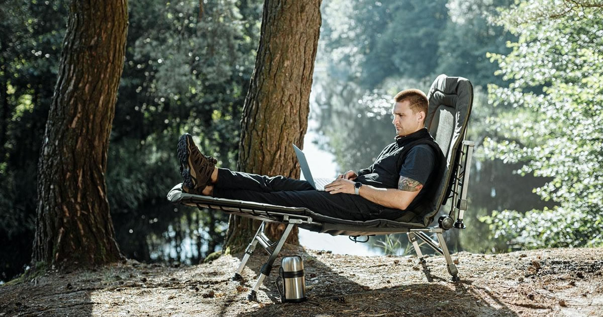 Man outdoors at summer camp lounging in chair writing on laptop