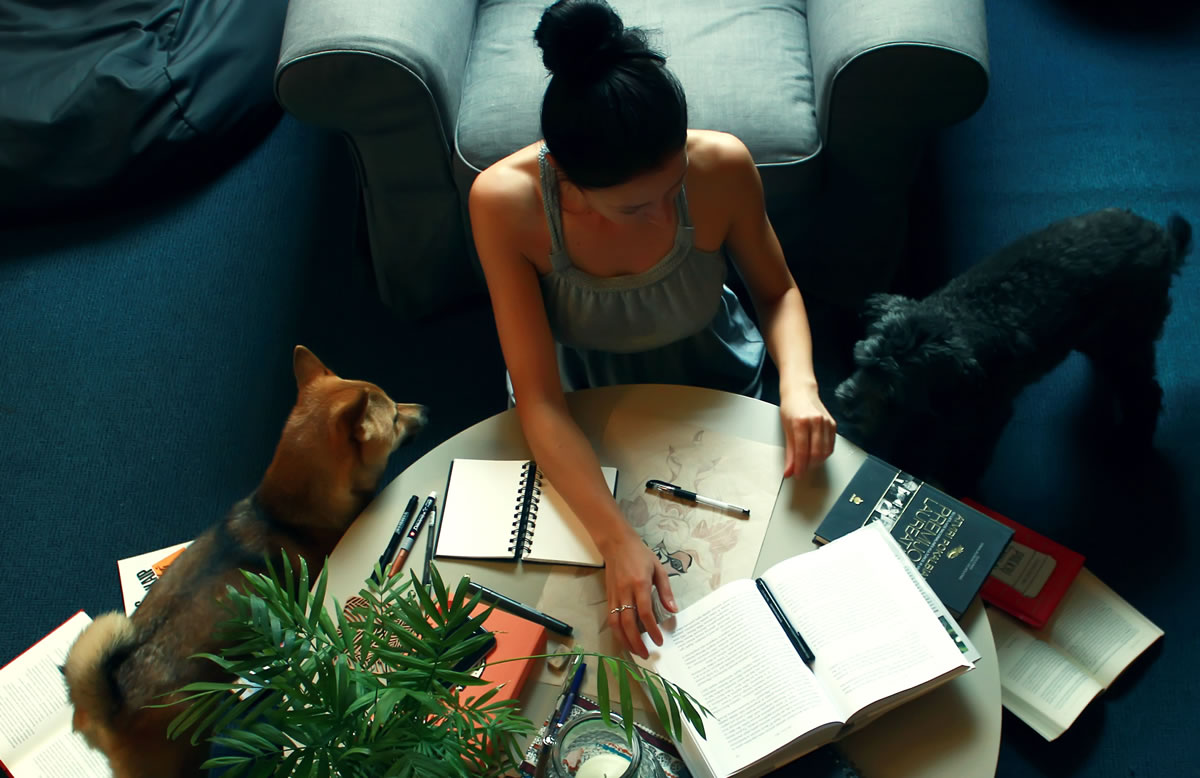 Overhead photo of woman sitting on floor at home between the couch and a coffee table with books, notebooks, and pens, with dogs nearby