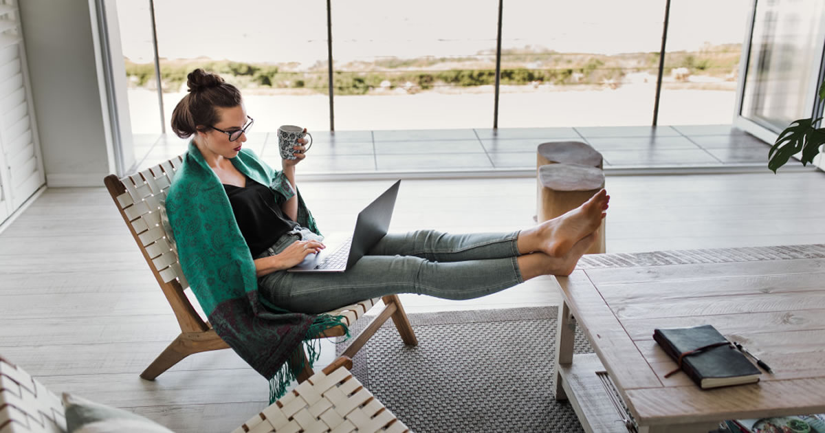 Women working on laptop at home while holding coffee and with her feet up on coffee table