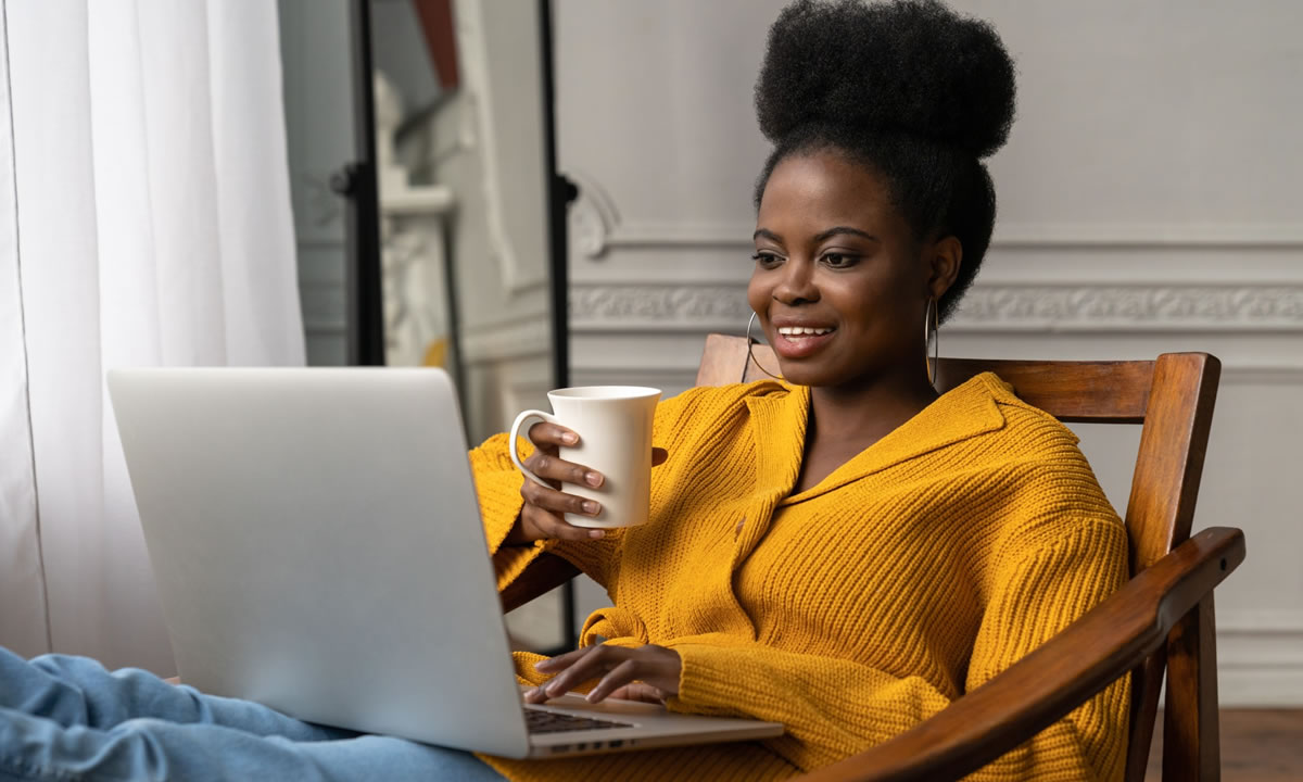 Smiling woman sitting in chair with legs up while using laptop and holding coffee mug