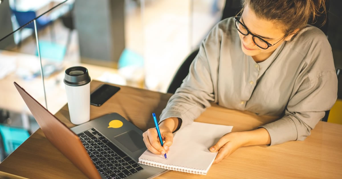 Woman at desk with laptop writing in notebook