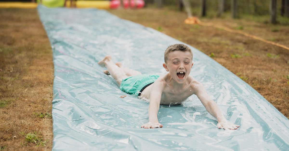 Happy young boy sliding down a water slippery slide on his stomach in a garden