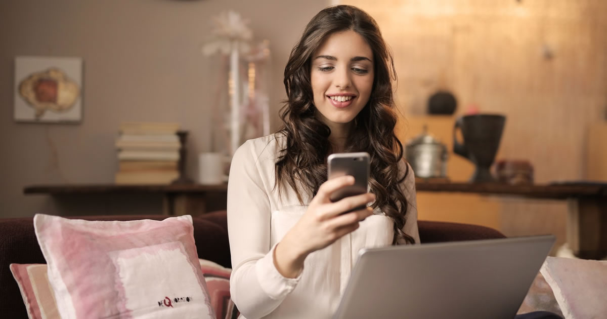 happy woman sitting on a sofa with laptop playing on smartphone