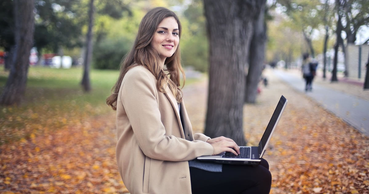 confident female freelancer writing on laptop in park in autumn