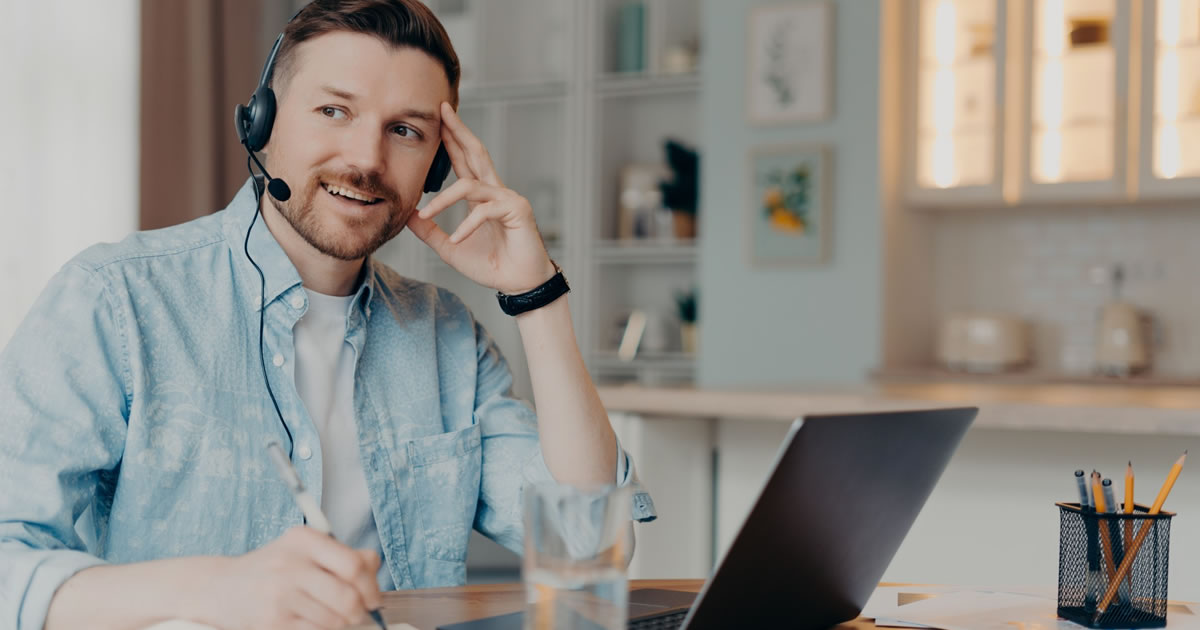 Thoughtful male writer makes notes uses laptop computer and headset and writes down notes in notepad