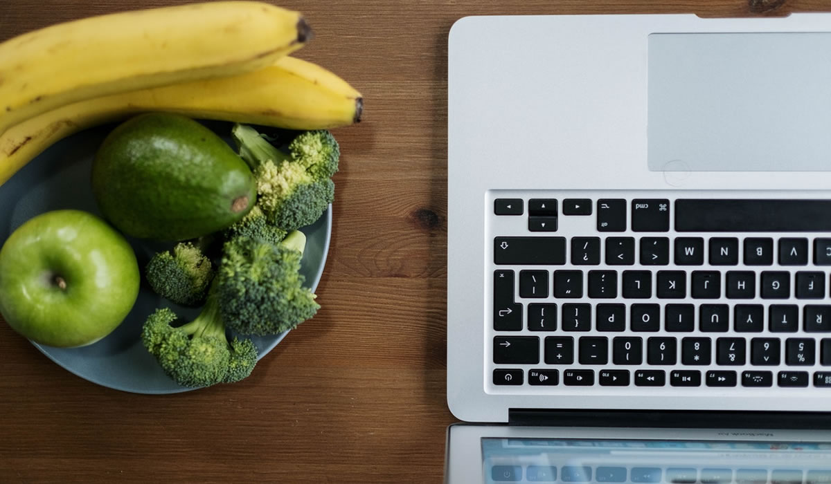 Laptop on table next to bowl of bananas and other fruits and vegetables