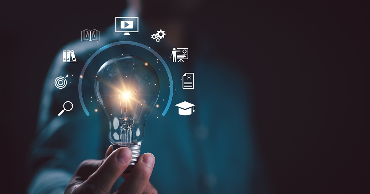Hand holding a lit light bulb against a dark background, with glowing icons hovering around it, including symbols for video, books, gears, teaching, documents, graduation, a target, magnifying glass, and folders, suggesting learning, ideas, and skill-building.