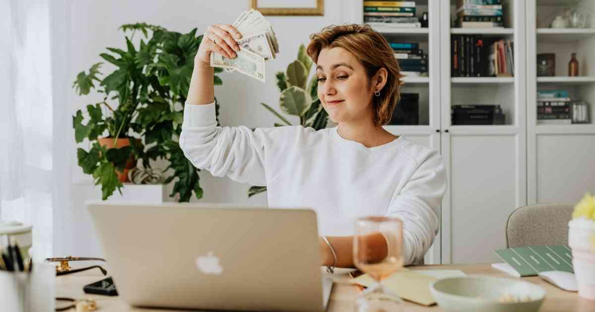 Woman sitting at a desk with a laptop, smiling confidently while fanning a stack of cash in one hand; she’s surrounded by plants, bookshelves, and work materials, suggesting successful remote or freelance work.