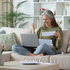 Woman sitting cross-legged on a couch at home, holding papers and celebrating with a raised fist while looking at a laptop and holding a check, showing excitement about a payment.