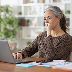 Woman setting at a table thinking, with a laptop, and a notebook and pen nearby, showing reflection and planning.